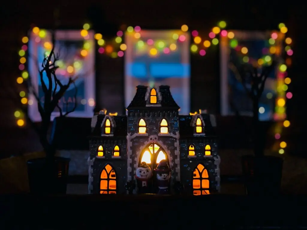 A small, decorative Halloween gingerbread house is illuminated with warm light from within, surrounded by two bare trees. Colorful blurred fairy lights twinkle in the background, creating a festive atmosphere.