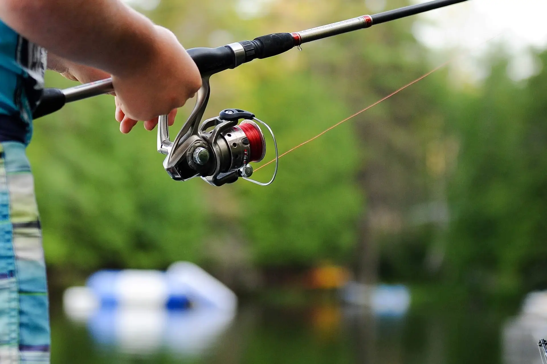 Close-up of a person holding a fishing rod with a red reel, enjoying free outdoor activities near a calm water body. The background is blurred with green trees and hints of a boat, suggesting a peaceful, outdoor setting.