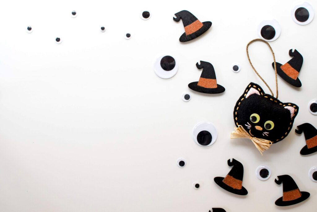 A Halloween-themed craft kit featuring a black cat ornament, witch hats, and googly eyes scattered on a white background. The cat ornament, adorned with facial features and a ribbon, comes with a string for hanging—perfect for your festive crafting fun!