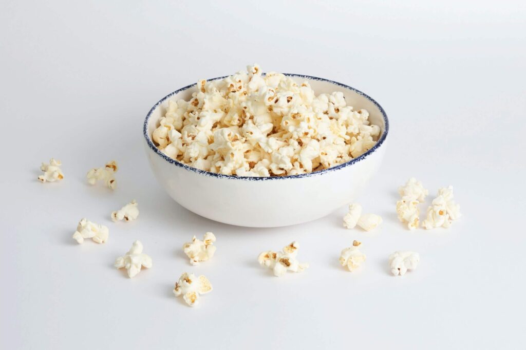 A white bowl filled with popcorn sits on a white surface, evoking cozy scenes perfect for back-to-school movie nights. Several popped kernels are scattered around the bowl, which is adorned with a thin, dark blue rim.