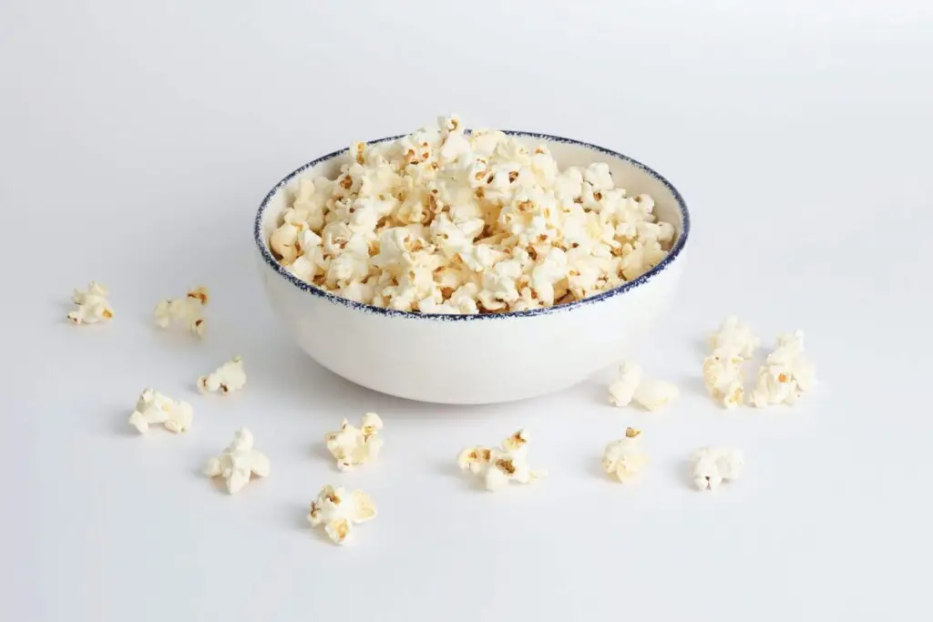 A white bowl filled with popcorn sits on a white surface, evoking cozy scenes perfect for back-to-school movie nights. Several popped kernels are scattered around the bowl, which is adorned with a thin, dark blue rim.