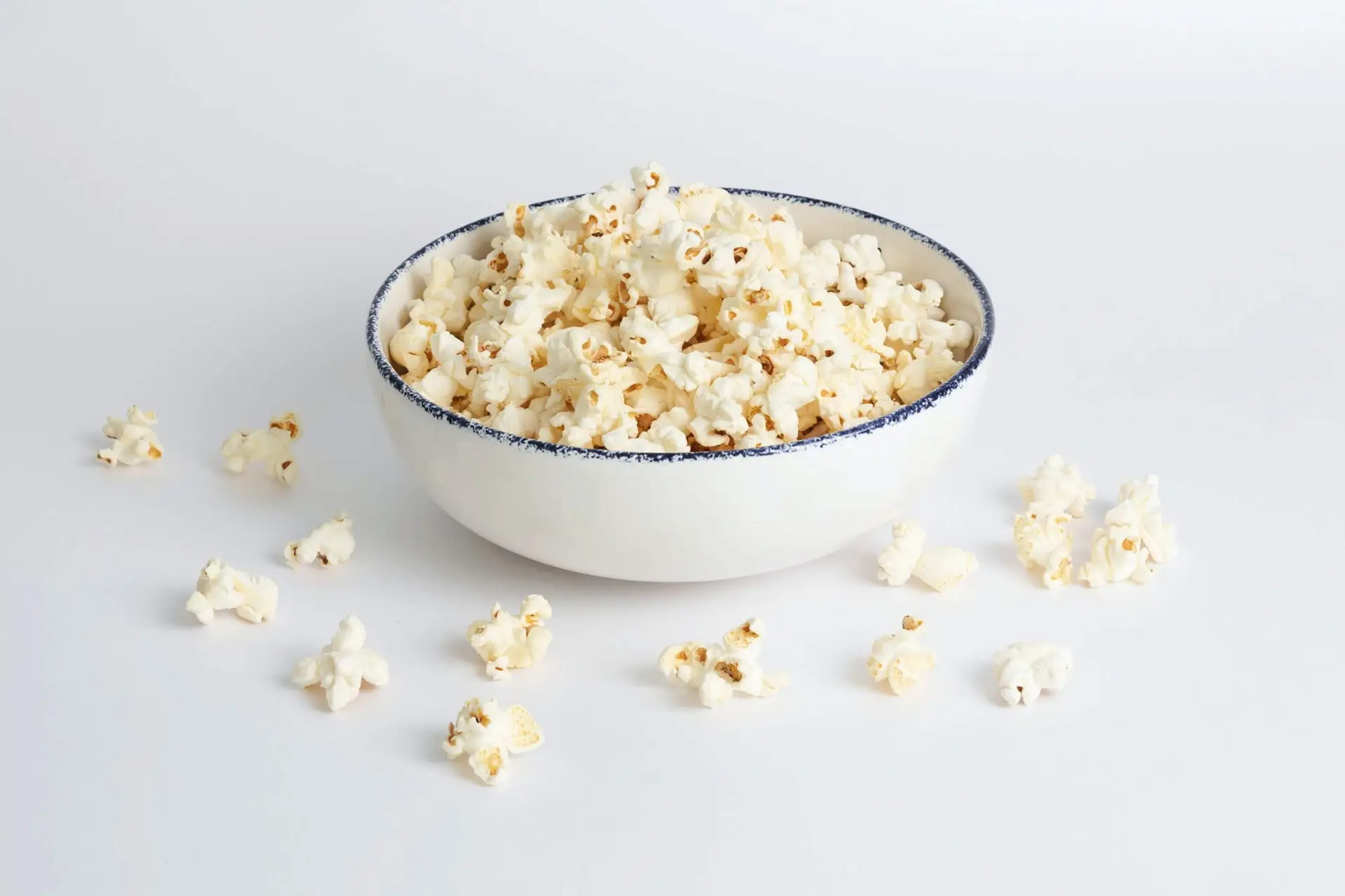 A white bowl filled with popcorn sits on a white surface, evoking cozy scenes perfect for back-to-school movie nights. Several popped kernels are scattered around the bowl, which is adorned with a thin, dark blue rim.