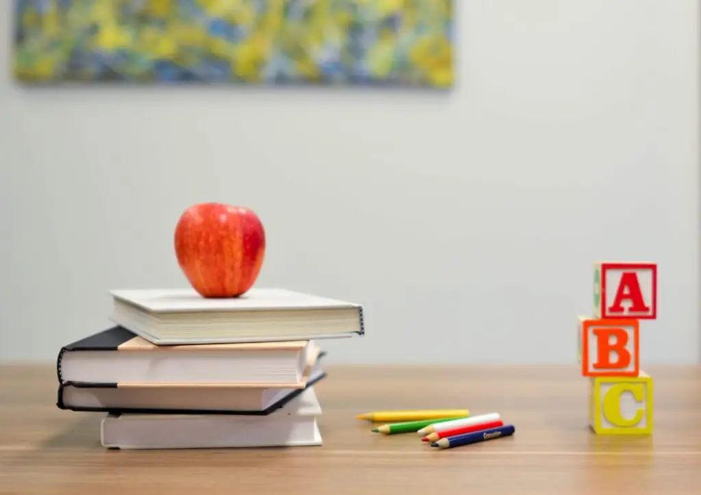 A stack of books with a red apple on top sits on a wooden table, perfect for back to school night. Nearby are colorful pencils and three alphabet blocks arranged to spell ABC. A blurred abstract painting adds a touch of creativity in the background.