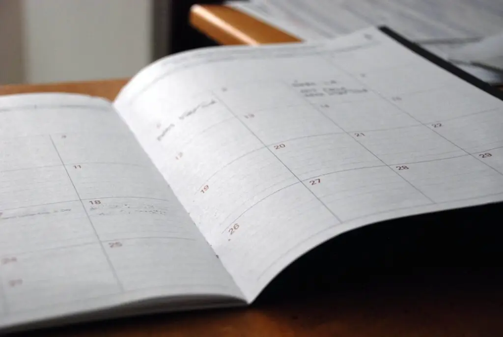 An open planner displays a monthly calendar with dates but no visible notes, resting on a wooden desk with some papers in the background. The focus is on the blank pages, eager to organize a summer schedule for kids or plan upcoming events.