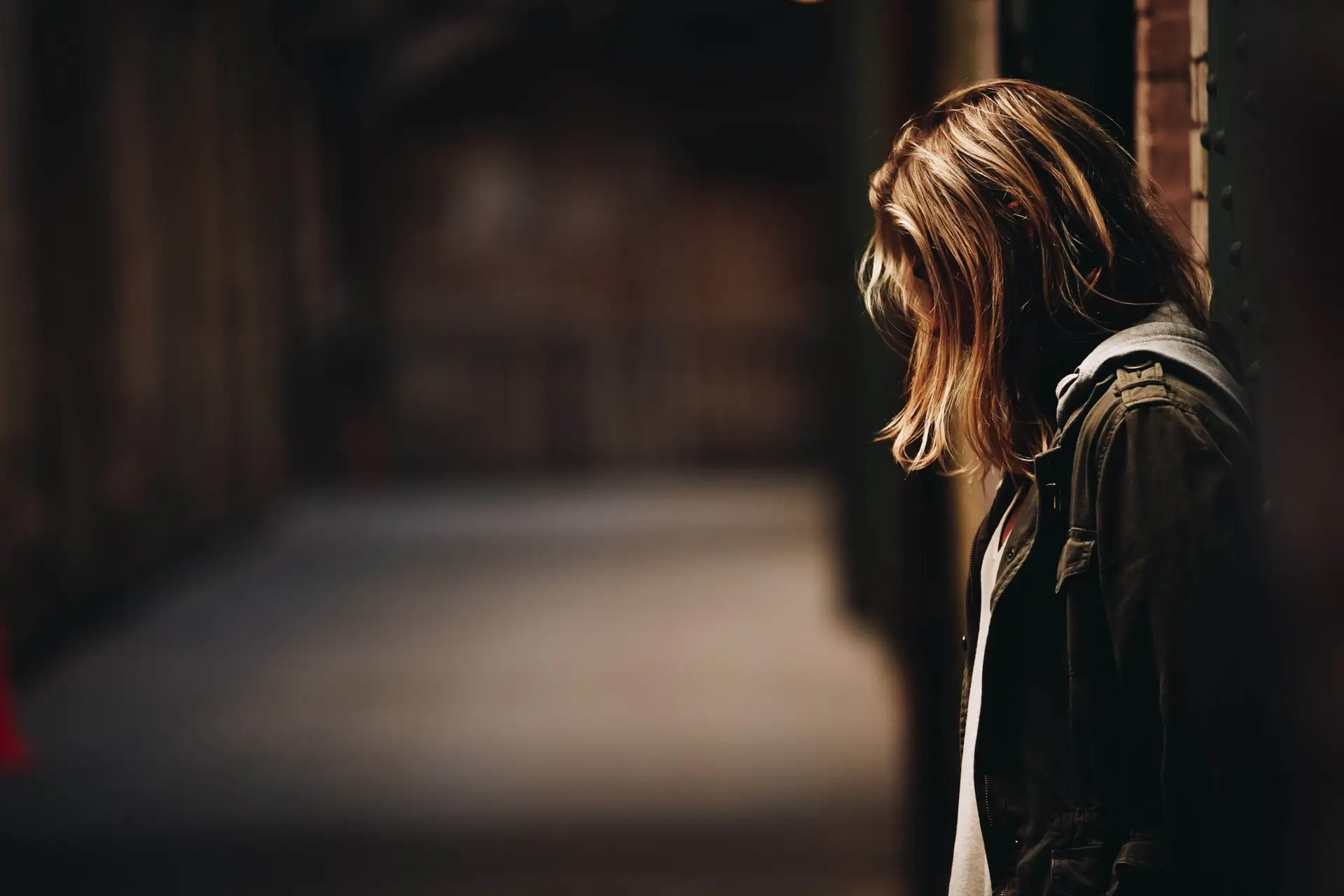 A person with shoulder-length blonde hair stands in a dimly lit hallway, leaning their head against a wall. They wear a dark jacket and face away from the camera, embodying the contemplative mood often felt when reflecting on how social media seriously harms your mental health.