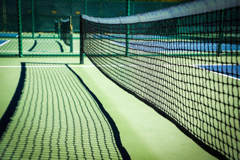 A close-up view of a tennis net casting a shadow on a sunlit court resembles the pattern often seen in games with strict elements like the rules for pickleball. The green surface contrasts with the dark net, while an out-of-focus net appears in the background.