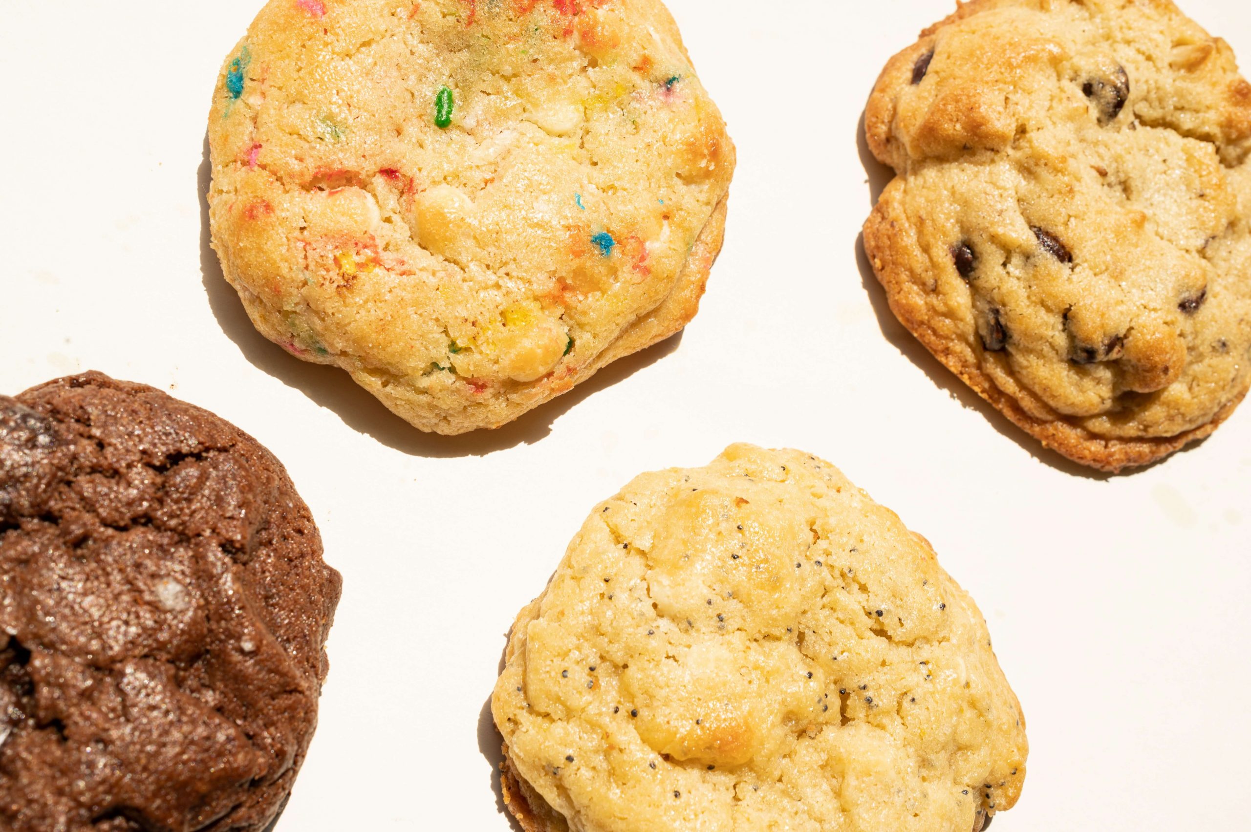 A close-up of four back-to-school cookies on a white surface. The assortment features a chocolate cookie, a colorful sprinkle cookie, a chocolate chip cookie, and a vanilla cookie—each golden-brown with a slightly crispy texture, perfect for those nostalgic school days.