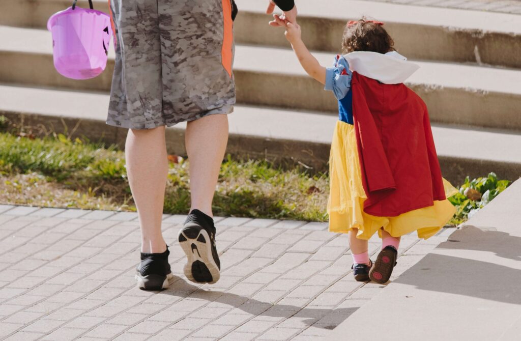 A child dressed in a Snow White costume, part of Disney family Halloween costumes, walks hand in hand with an adult wearing camouflage shorts. The child carries a purple Halloween bucket as they stroll outdoors on a sunny day.