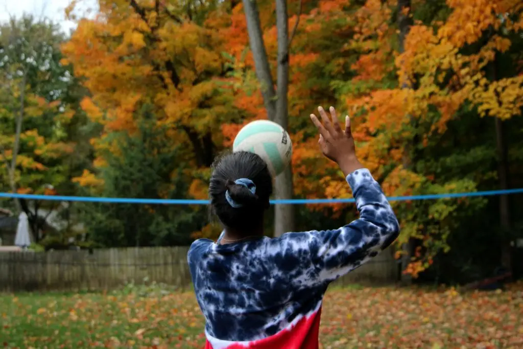 A person wearing a tie-dye sweatshirt prepares to hit a volleyball over the net, embodying the excitement of fall sports for kids. The background bursts with trees adorned in vibrant autumn leaves, ranging from green to orange.