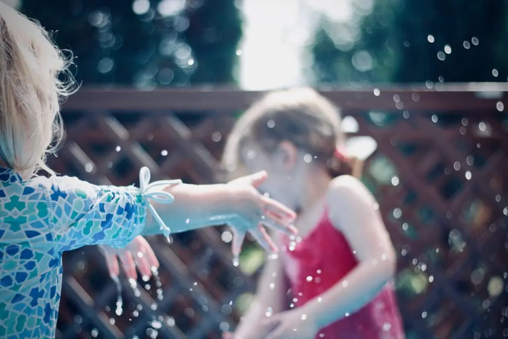 Two children delight in water games for kids on a sunny day. One, in a patterned blue outfit, reaches out joyfully while the other, wearing a red shirt, is partially blurred in the background. Water droplets are suspended in the air around them.