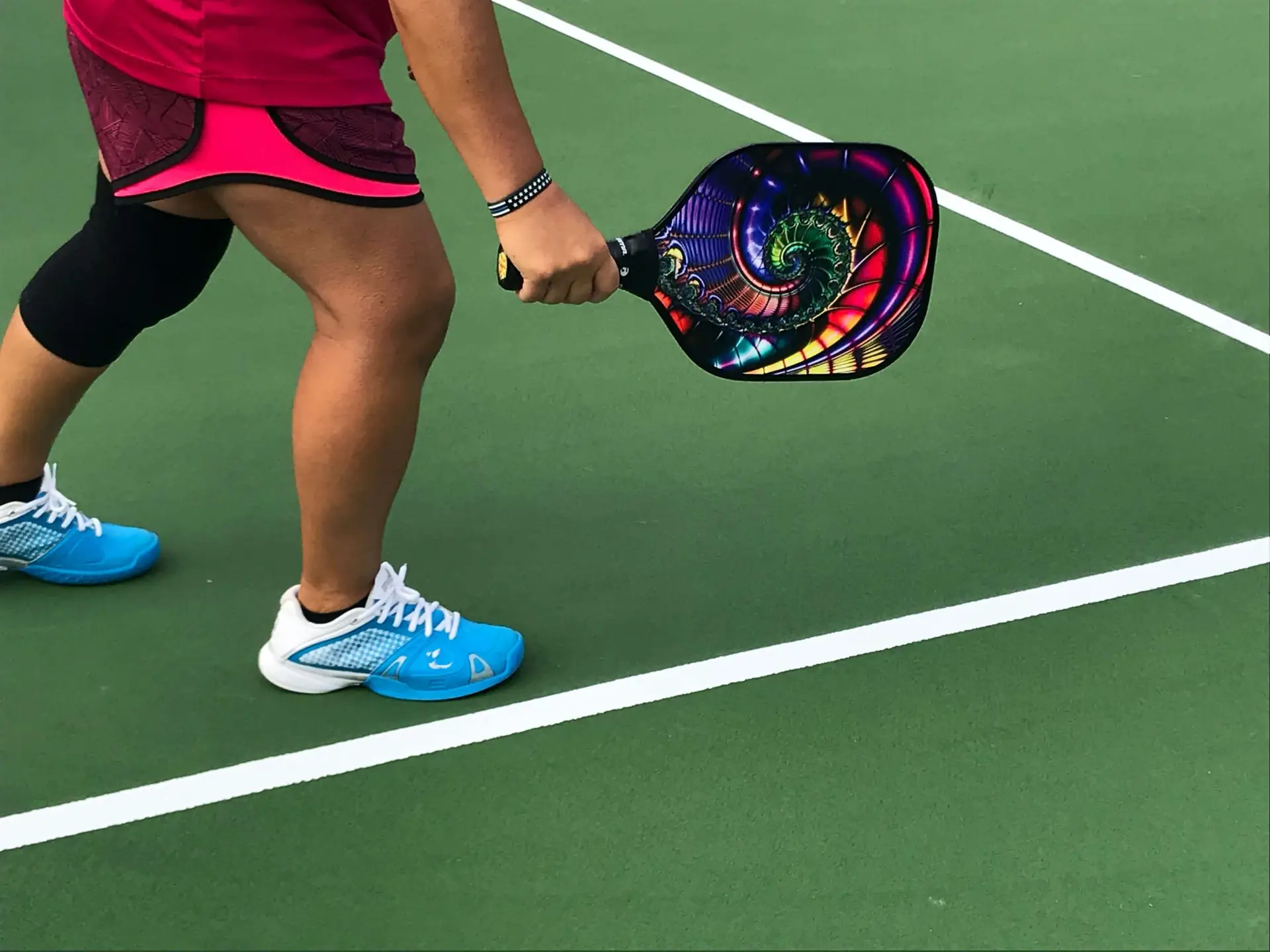 A person grips one of those vibrant pickleball paddles on a green court, clad in a red sports outfit and blue sneakers, knee brace ready. The colorful paddle boasts a mesmerizing spiral design as they prepare to dive into the lively game of pickleball.