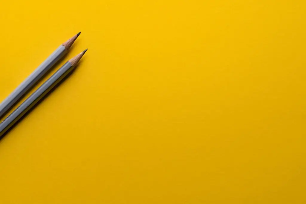 Two sharpened pencils with gray bodies are placed diagonally on the left side of a bright yellow background, crafting a simple and minimalistic composition that's perfect for catching the eye—and bringing back to school jokes to mind. The ample negative space adds to its charm.