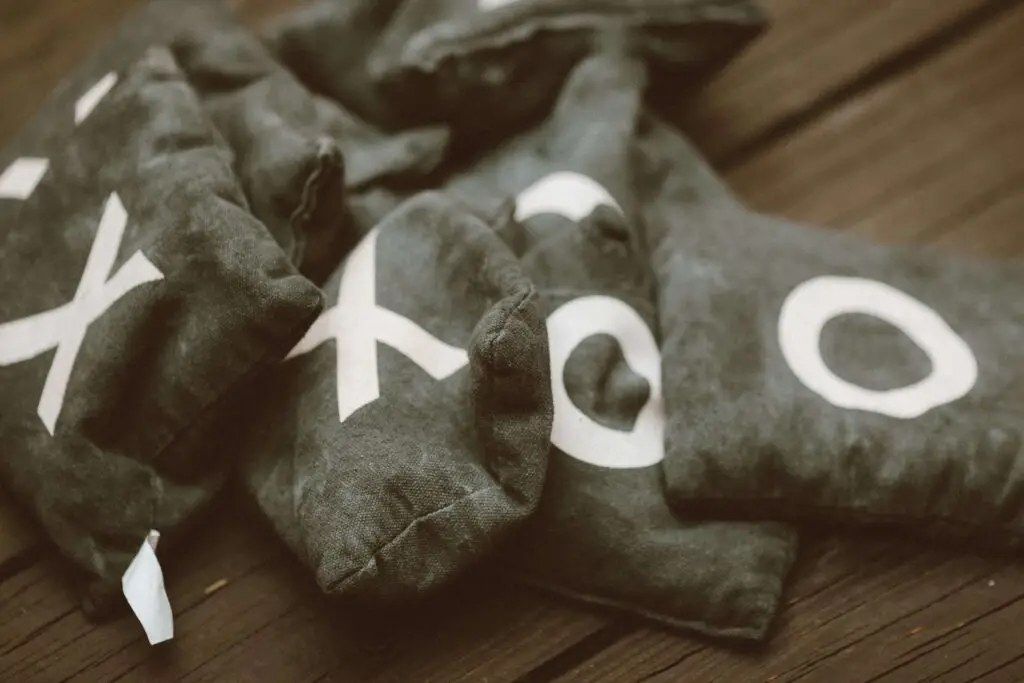 A pile of dark fabric bean bags featuring white X and O symbols, reminiscent of a giant kids cornhole game, rests on a wooden surface.