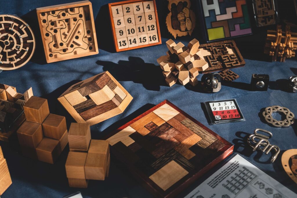 A collection of wooden and metal puzzles, including 3D shapes, maze puzzles, and number grids, displayed on a blue surface. Sunlight casts shadows like mystery games for kids, highlighting the intricate designs and craftsmanship of each puzzle piece.