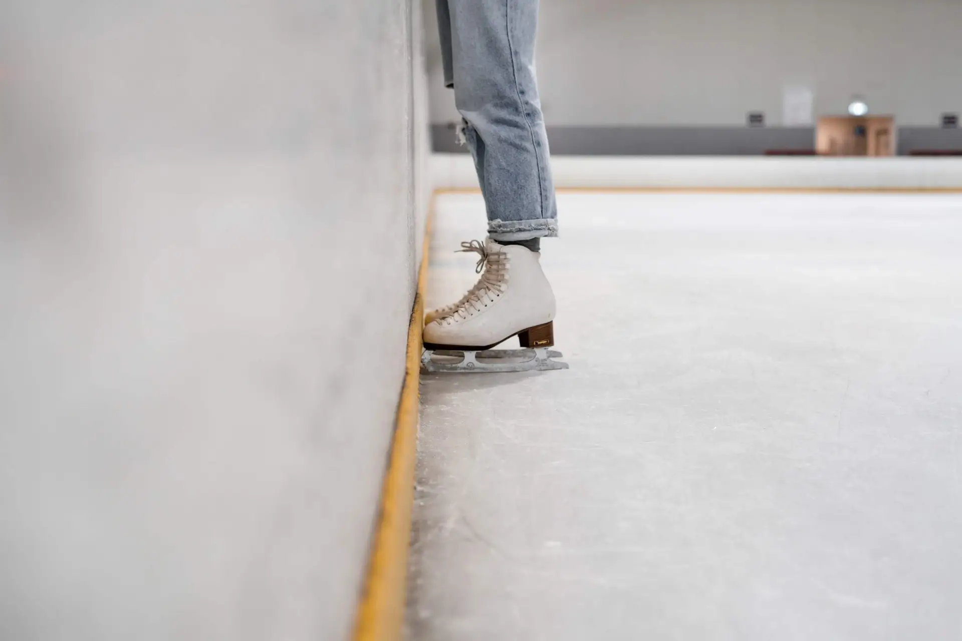 Close-up of a person wearing white ice skates, gently touching their blade against the wall of an ice rink. Their rolled-up jeans hint at a casual vibe, embodying the essence of winter sports for kids. The rink appears empty and well-lit, inviting young skaters to explore its gleaming surface.