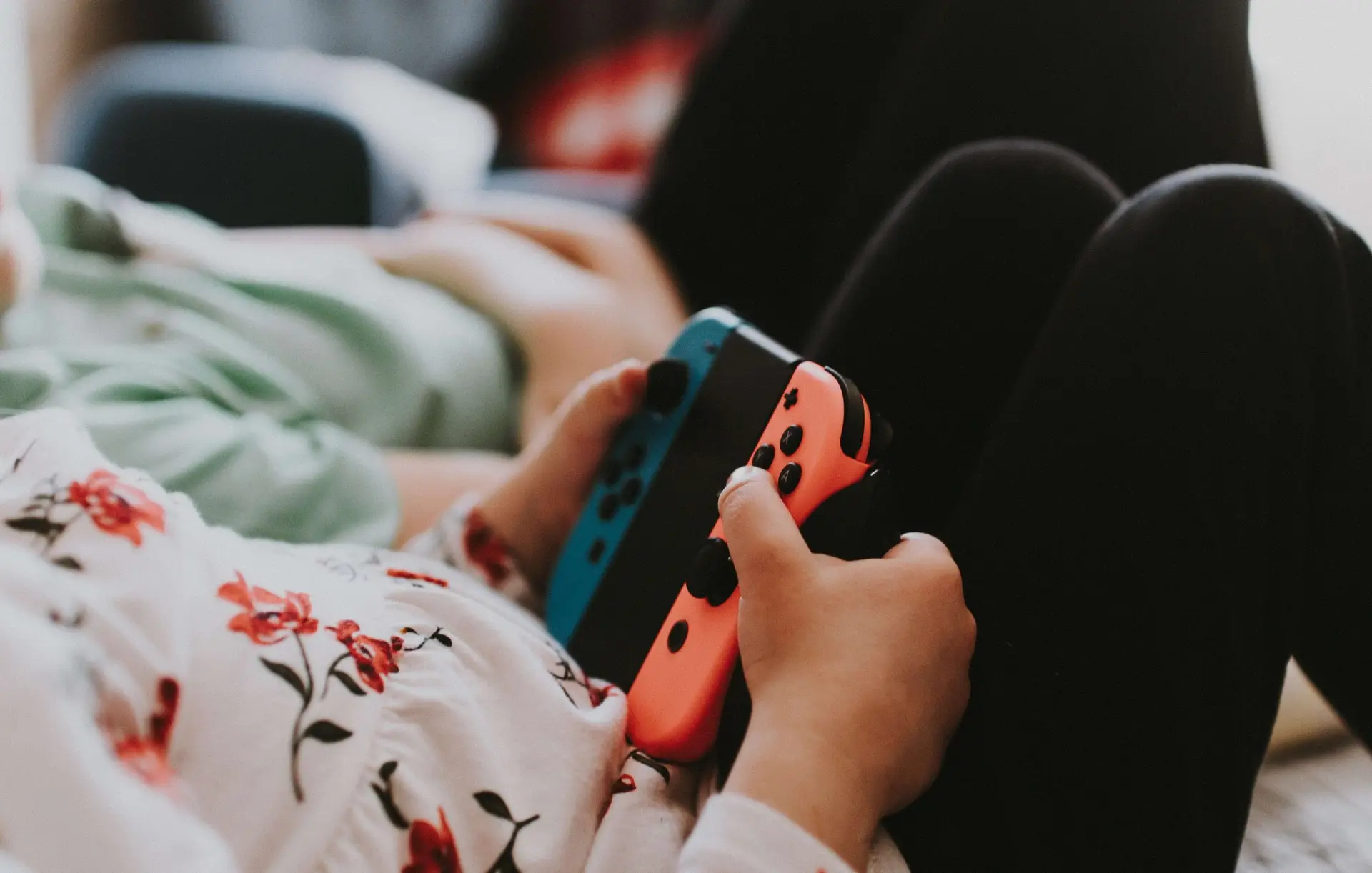 Two people are seated, holding video game controllers with a focus on one person's hands. They wear a floral-patterned shirt, and red and blue controllers are visible. The scene captures the essence of a relaxed video games session.