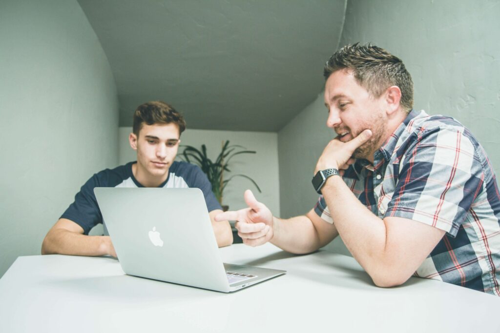 Two men sit at a table, immersed in a discussion about parenting teens and tweens. One gestures toward a silver laptop in front of them, emphasizing his point. The setting is modern and minimalist, with a plant adding a touch of nature to the background.