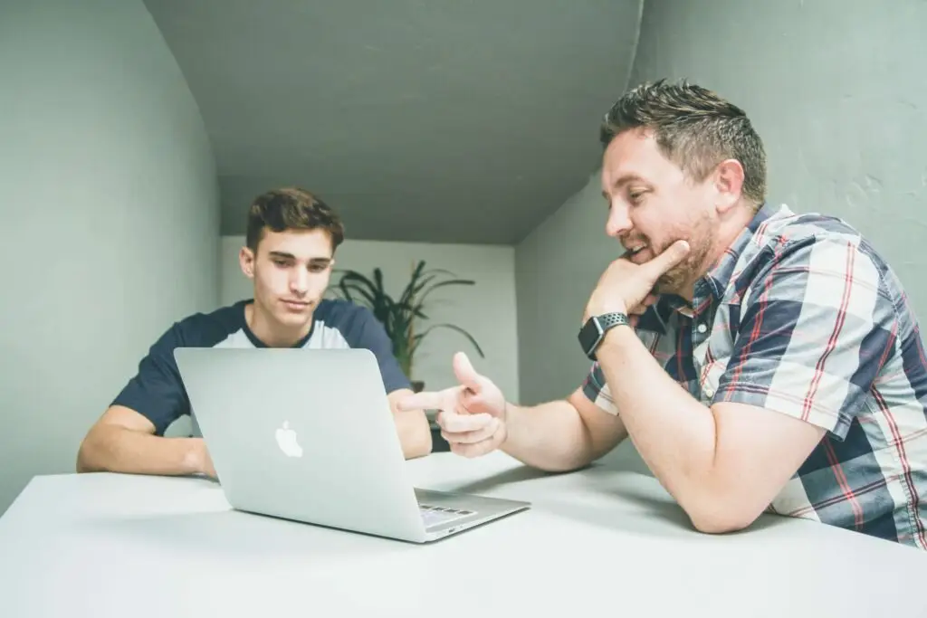 Two men sit at a table, immersed in a discussion about parenting teens and tweens. One gestures toward a silver laptop in front of them, emphasizing his point. The setting is modern and minimalist, with a plant adding a touch of nature to the background.