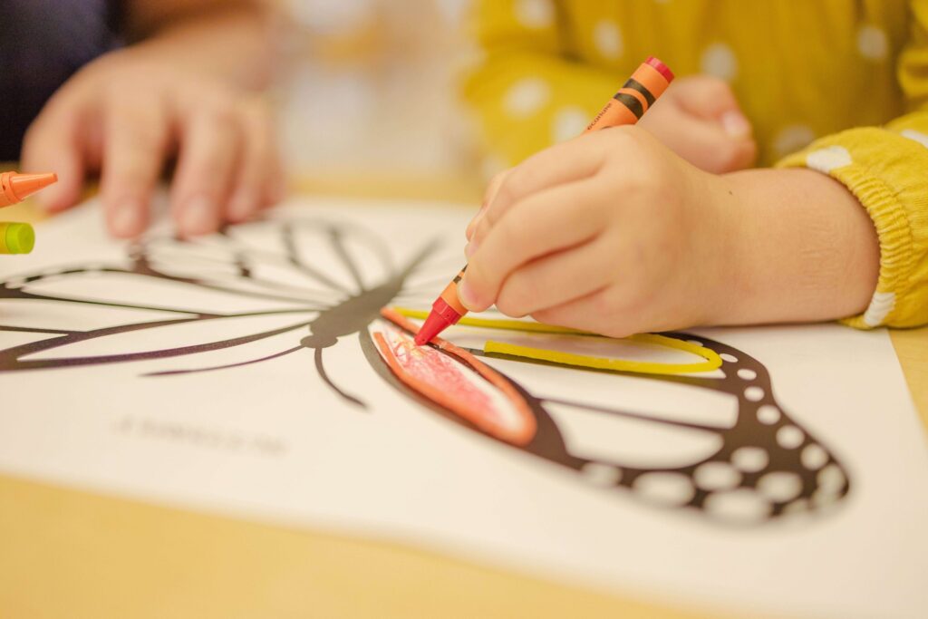 A child colors a butterfly illustration with orange and yellow crayons, engaging in enrichment activities for kids. The butterfly has bold, black outlines, and the child wears a yellow polka dot shirt. An adult's hand is partially visible assisting. The scene is bright and focused on creativity.