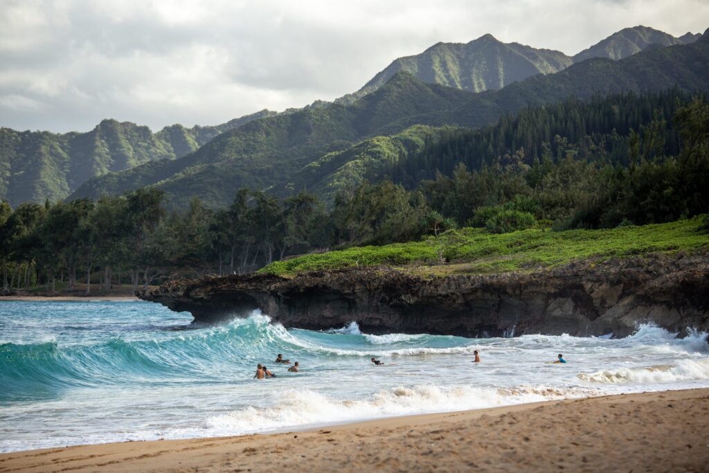 A scenic Hawaii beach with several people swimming in the ocean offers a tranquil escape. Lush green mountains and dense forest serve as the backdrop under a cloudy sky, while gentle waves crash on the sandy shore, making it a natural paradise worthy of any travel tips.