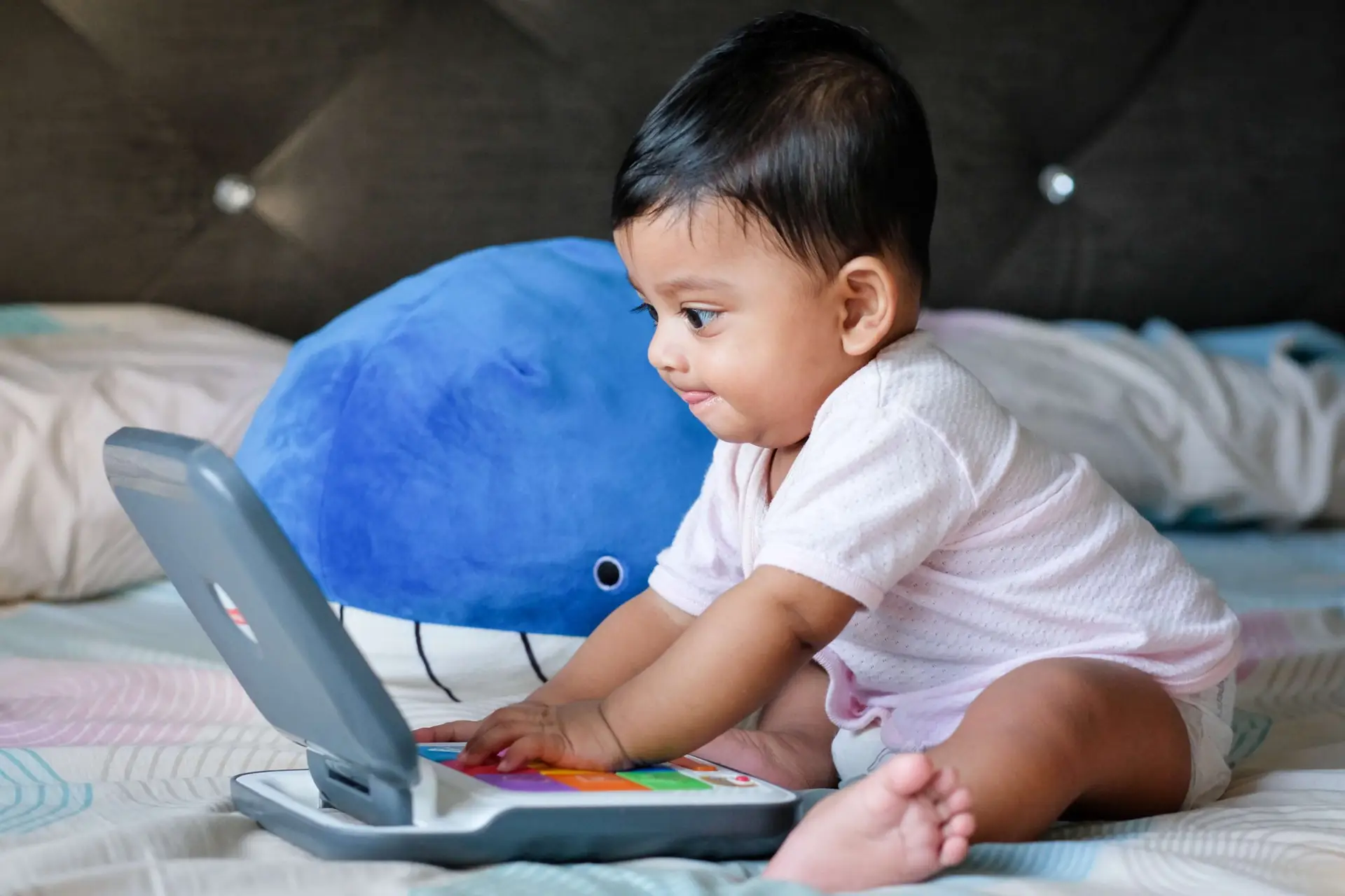 A baby in a pink outfit sits on a bed, deeply engaged with a toy laptop, resembling someone immersed in a kids' wordle game. A blue whale-shaped pillow playfully adds to the charming scene.