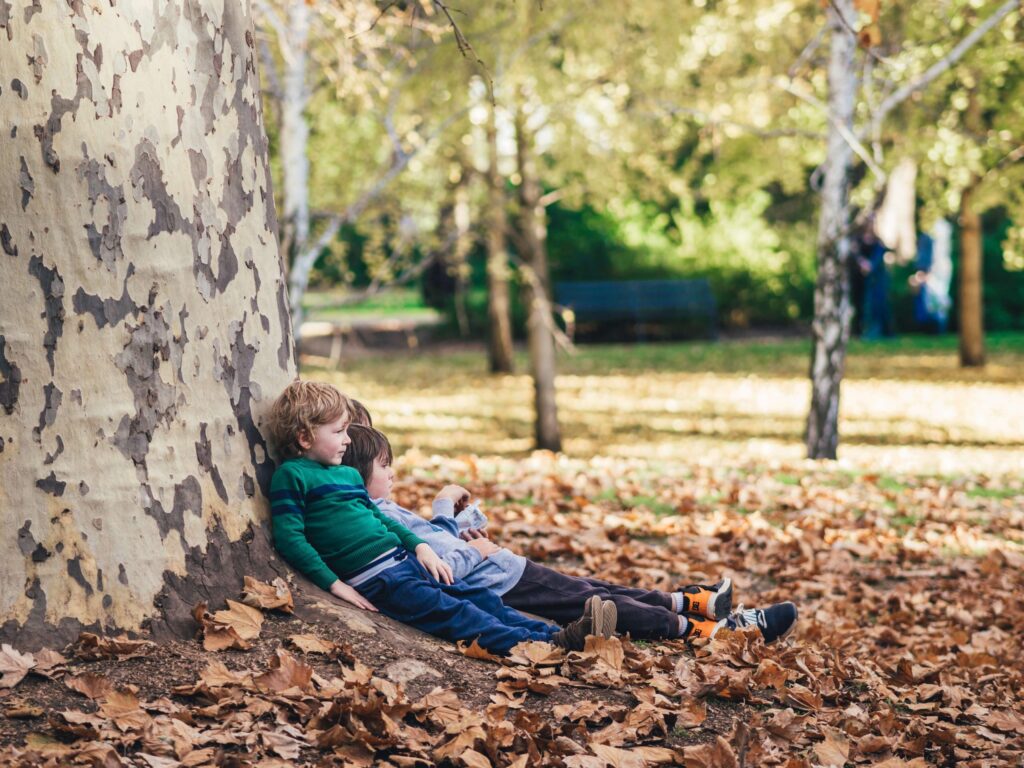 Three children sit against a large tree trunk, surrounded by vibrant fallen leaves in a park, embodying the perfect fall activity for preschoolers. They appear relaxed, enjoying the sunny day under the trees. In the background, more trees and faint figures can be seen.