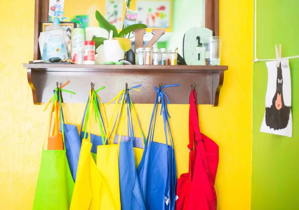 Colorful aprons hang on hooks below a shelf filled with various items, including jars, plants, and decorative letters against a bright yellow wall. A black-and-white rabbit illustration peeks out on the right, inspiring whimsical back-to-school crafts.