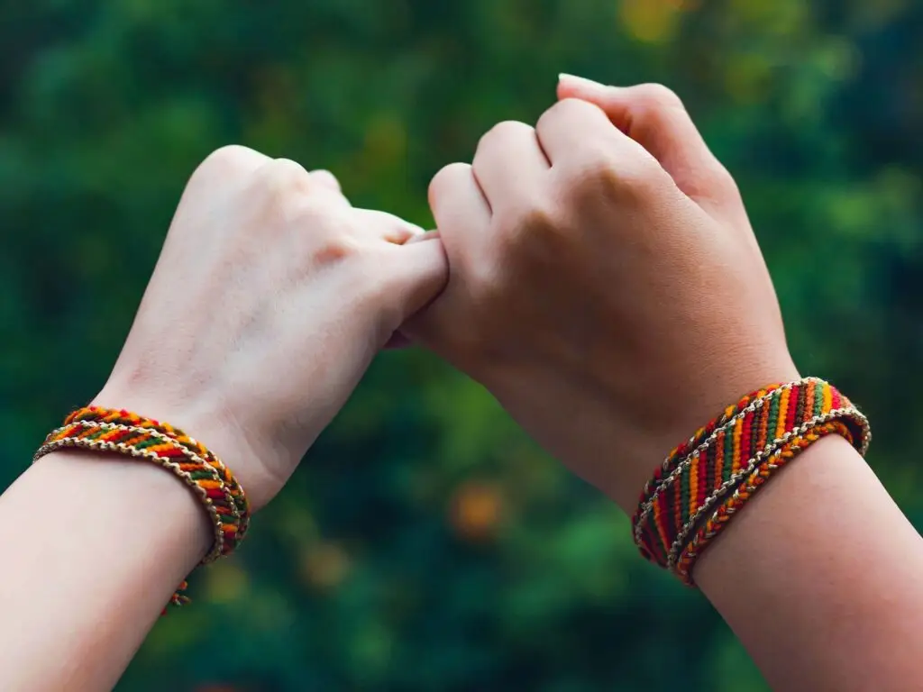 Two hands adorned with vibrant friendship bracelets make a pinky promise against a blurred green backdrop. The left sports a red and white design, while the right showcases red, yellow, and orange. These colorful threads carry the rich history of friendship bracelets, symbolizing trust and unity.