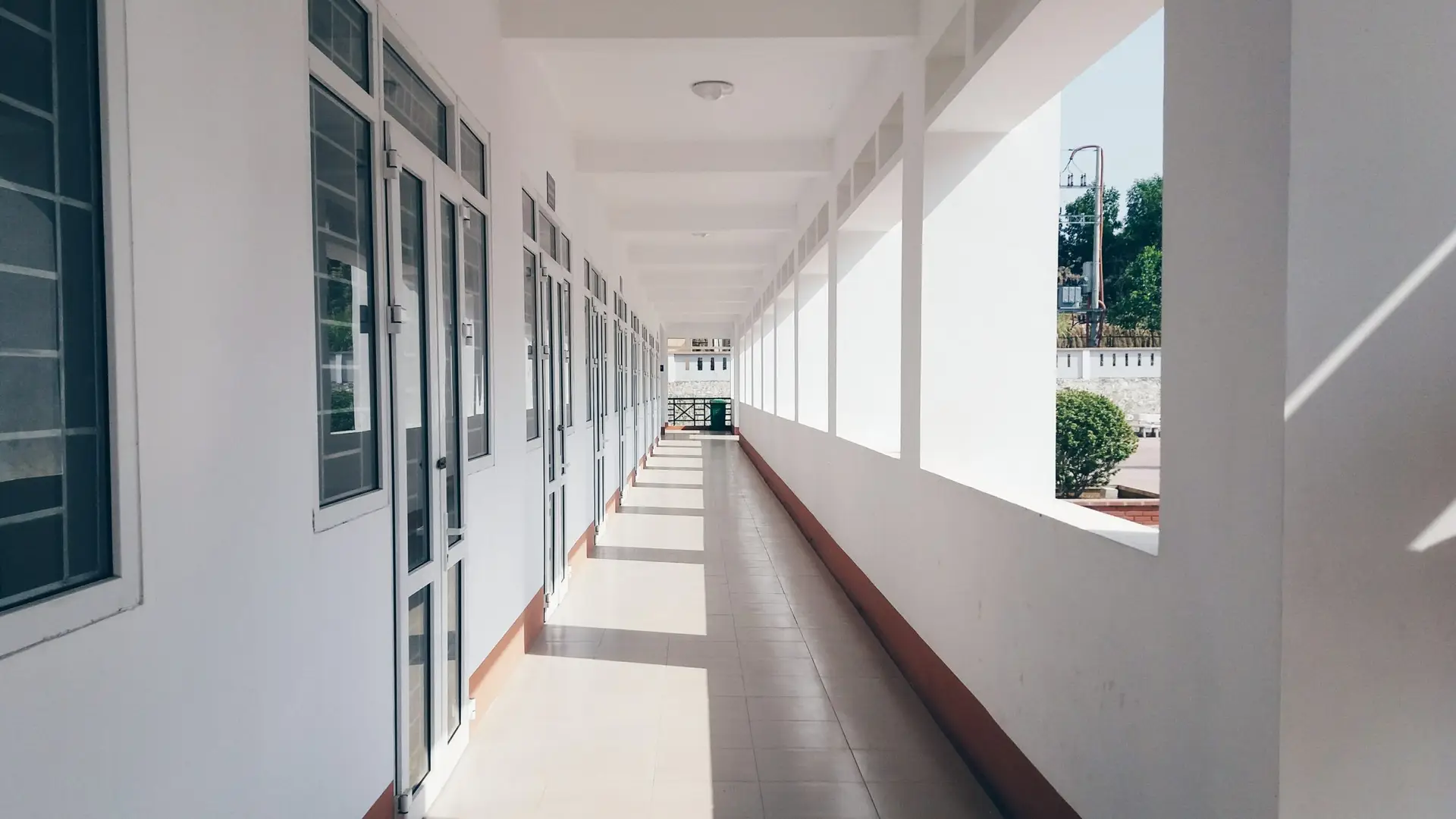 A bright, uncovered corridor with white walls and tiled floors. Large windows line the left side, while an open-sided railing allows sunlight to cast shadows on the right. As students meander, pondering why kids hate school, green foliage is visible at the end of the corridor.