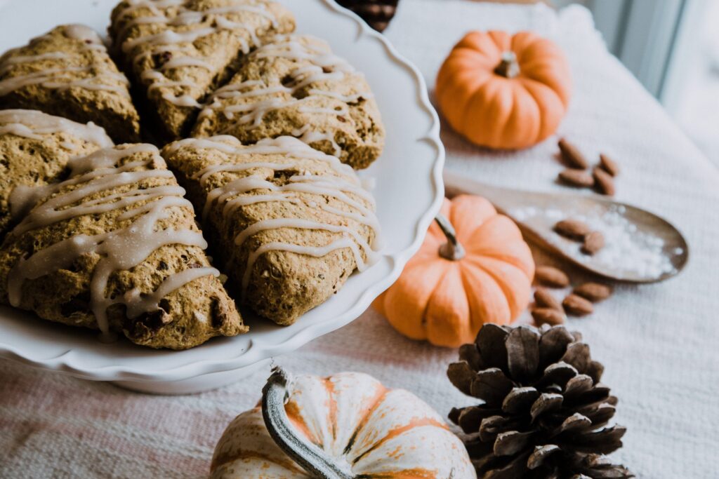 A plate of iced pumpkin scones, perfect fall snacks for kids, sits on a white cake stand surrounded by small decorative pumpkins, almonds, a wooden spoon filled with salt, and a pinecone on a white textured cloth.