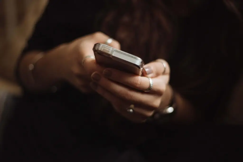 A person with dark hair, reminiscent of a stealthy predator, is holding a smartphone with both hands. Rings adorn their fingers, glinting subtly in the dim light. The focus is entirely on the hands and phone, capturing the intensity of their seemingly tactical grip.