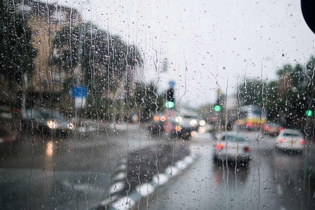 View through a rain-soaked window showing a busy urban street, reminiscent of rainy day movies. Blurry cars navigate the wet road beneath overcast skies. Traffic lights glow green, and the scene is softened by droplets on the glass.