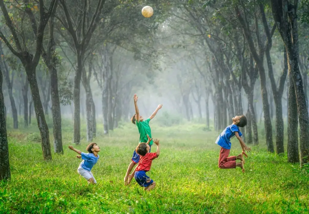 Four children are joyfully playing with a ball in a lush, green forest, embracing family exercise. They jump and reach for the ball amongst tall, dense trees, under a misty, overcast sky. The scene is vibrant and full of energy.