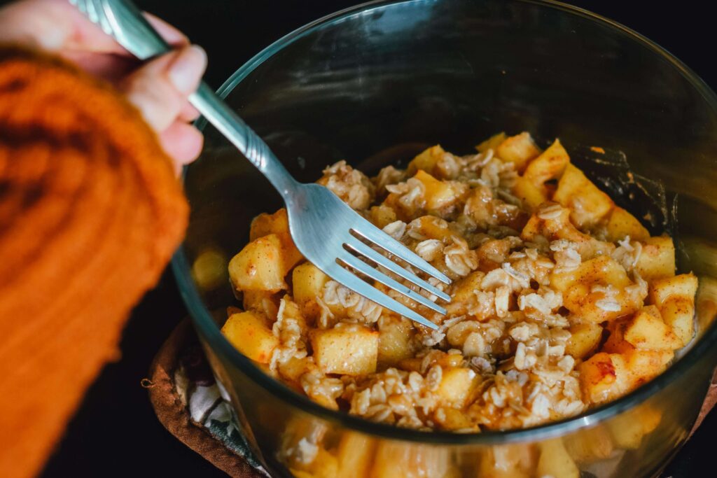 A person in an orange sweater stirs a gluten-free mixture of chopped apples, oats, and cinnamon in a glass bowl with a fork. The ingredients hint at a delicious fall dessert or breakfast dish.