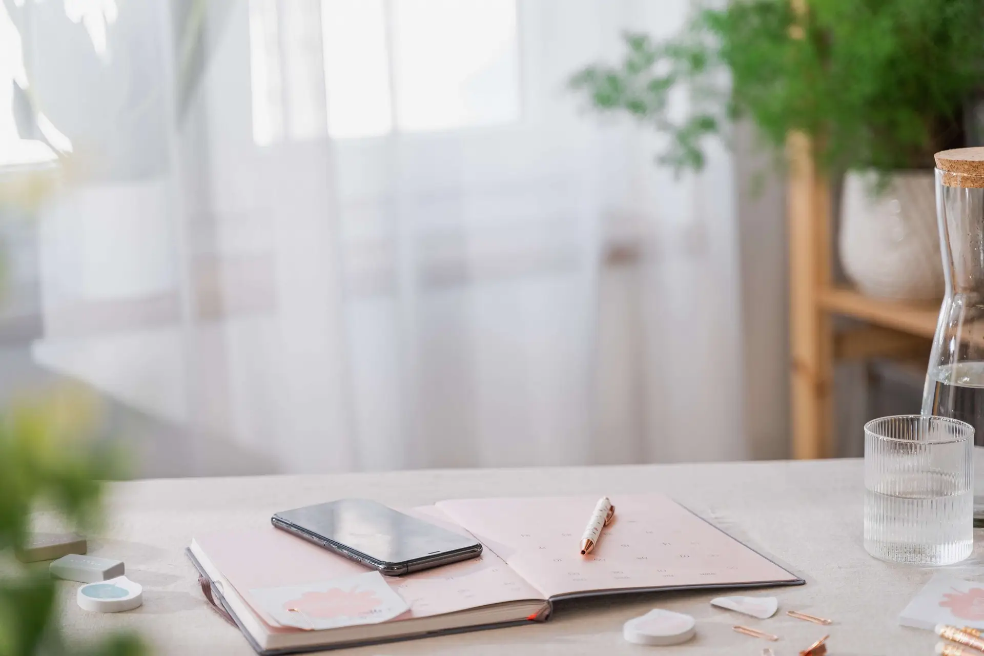 Open notebook on a table with a smartphone on top, beside a pen—a DIY chore chart in the making. A glass of water is nearby, and leafy plants are in the background. Natural light filters through sheer curtains, creating a serene atmosphere.