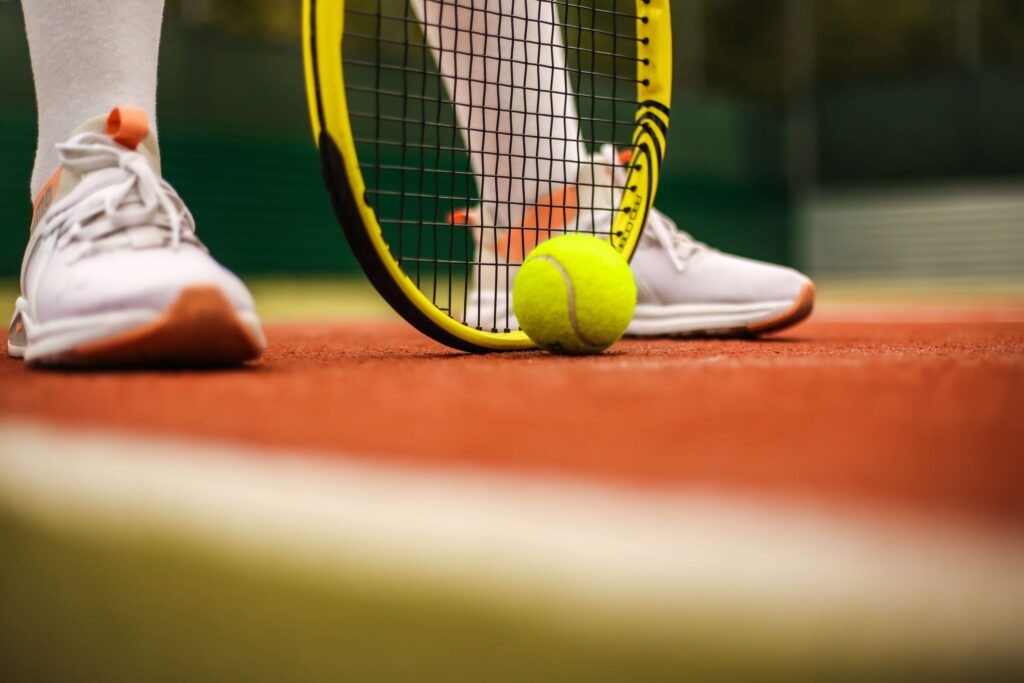 Close-up of a tennis player standing on a clay court, embodying the spirit of spring sports for kids with white sneakers accented in orange. A tennis racket stands upright beside a bright yellow ball, ready to inspire young athletes.