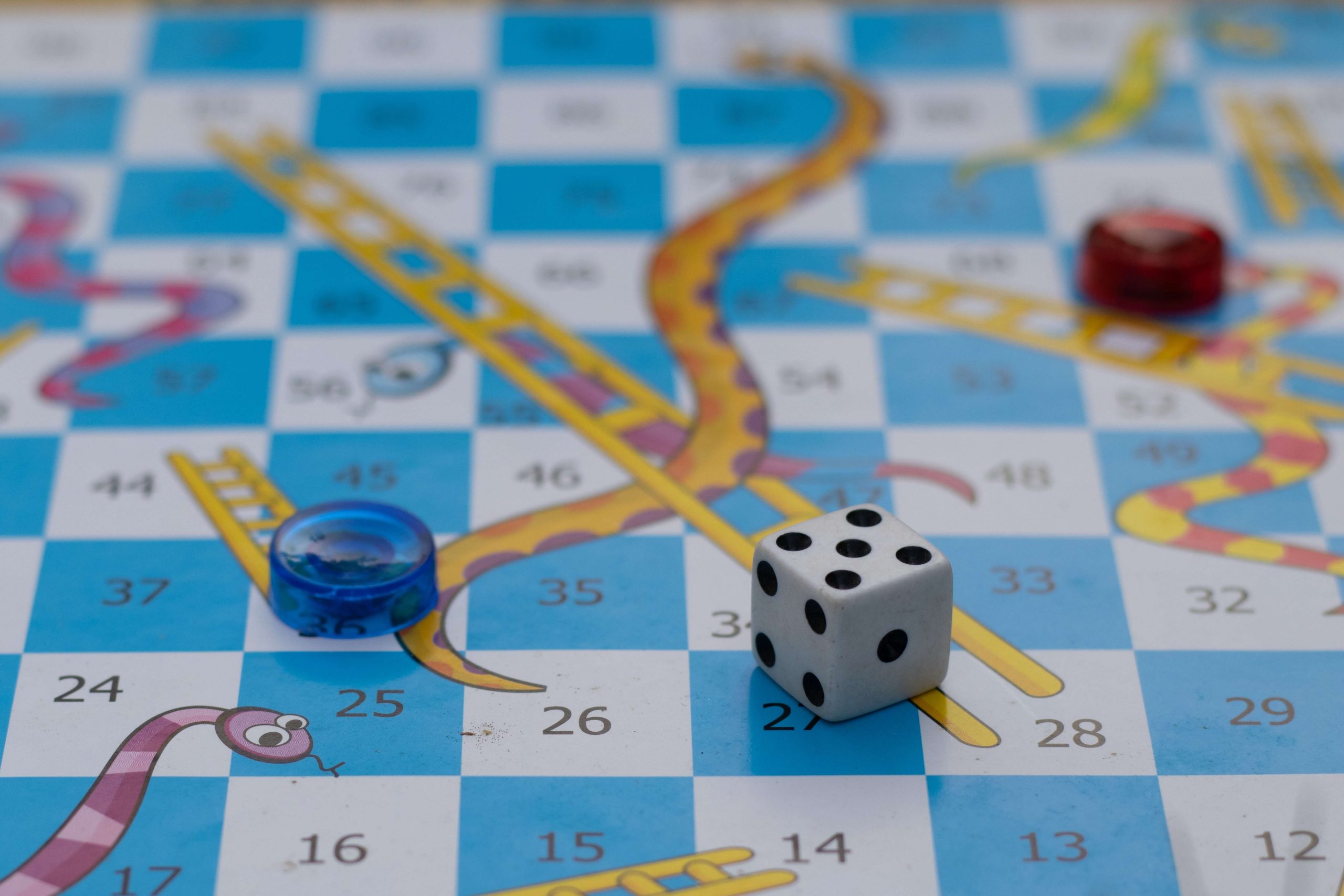 A perfect family game night idea: Snakes and Ladders with a white die showing four, alongside red and blue game pieces on a blue and white checkered board featuring illustrated snakes and ladders.