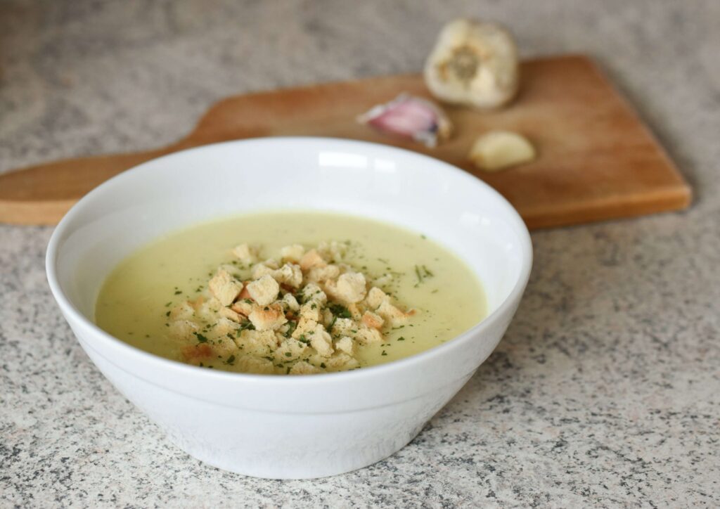 On a speckled countertop, a white bowl brimming with creamy soup, adorned with croutons and herbs, invites comfort. In the background, a wooden cutting board holds garlic bulbs and cloves—a perfect setting for Rainy Day Recipes that warm the soul.