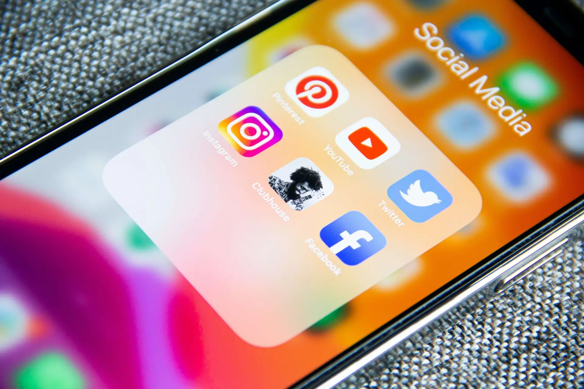 A smartphone screen displays a folder labeled Social Media with six app icons: Pinterest, Instagram, Clubhouse, YouTube, Twitter, and Facebook—tools that explain why kids are often addicted to social media. The phone lies on a textured surface, showing a colorful background.