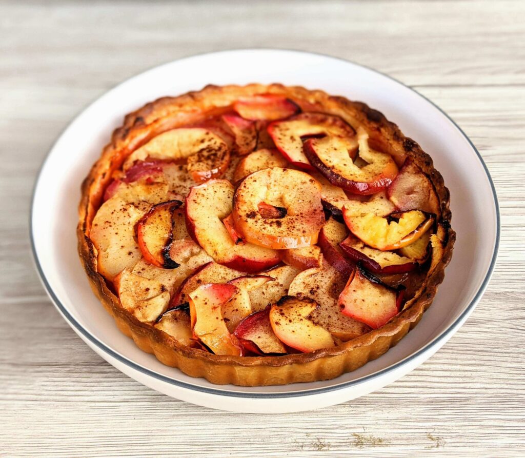 A freshly baked apple tart, embodying the essence of apple desserts for fall, is displayed on a white plate. The tart is golden brown with neatly arranged apple slices on top, lightly dusted with cinnamon. It sits on a light wooden surface, emphasizing a rustic presentation.