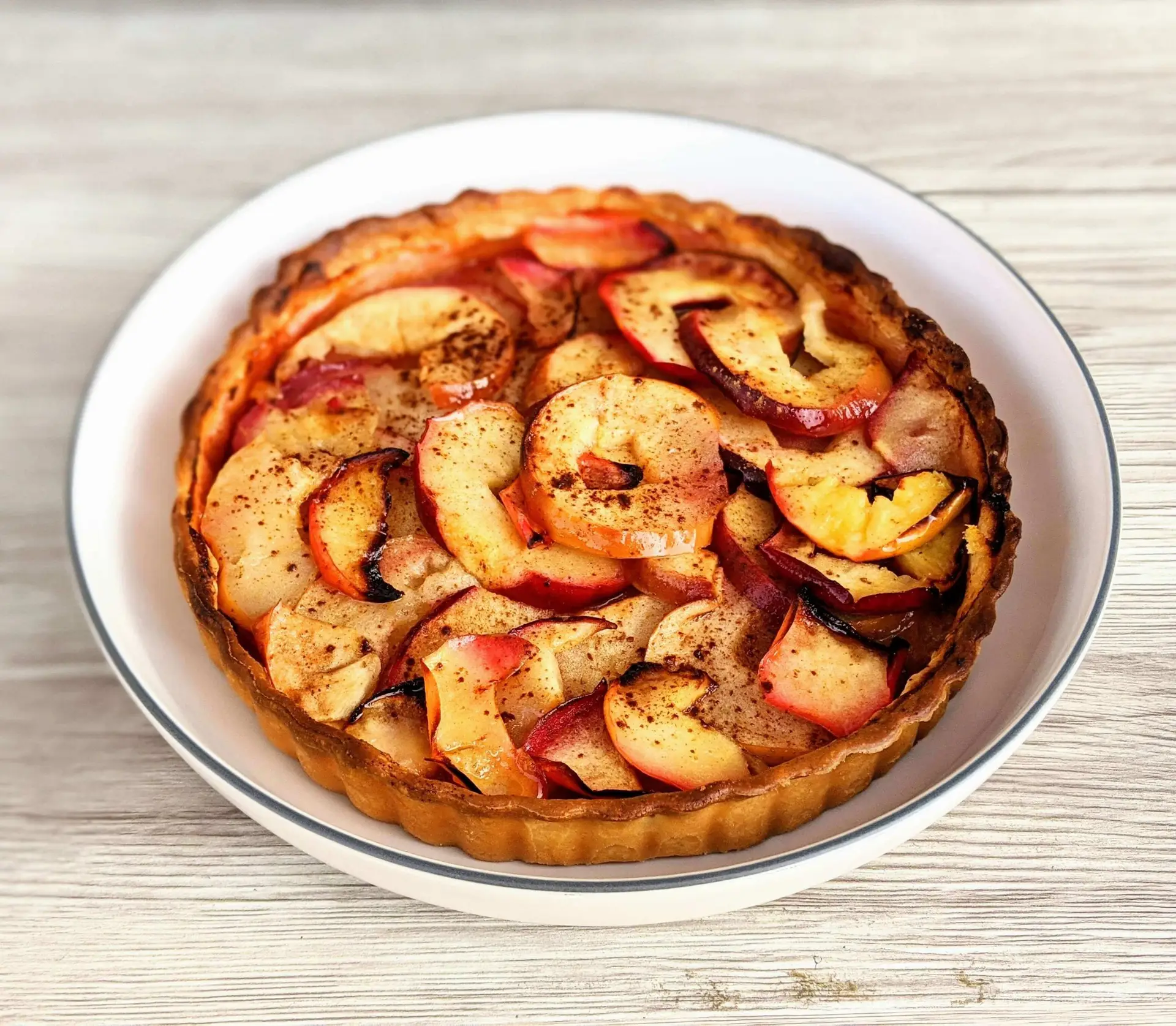 A freshly baked apple tart, embodying the essence of apple desserts for fall, is displayed on a white plate. The tart is golden brown with neatly arranged apple slices on top, lightly dusted with cinnamon. It sits on a light wooden surface, emphasizing a rustic presentation.