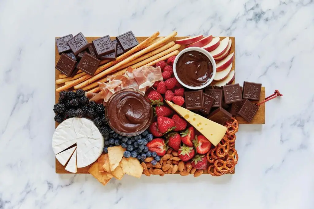 A Halloween Dessert Charcuterie Board features dark chocolate squares, breadsticks, apple slices, prosciutto, strawberries, blueberries, blackberries, almonds, pretzels, brie cheese, Swiss cheese, and chocolate dips elegantly arranged on a marble surface.