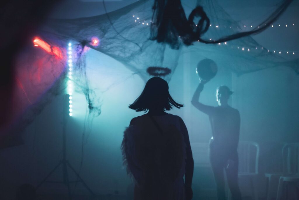 A dimly lit room with silhouetted figures, one wearing angel wings and a halo, and another holding a balloon. The space, adorned with cobwebs and colorful lights, evokes Halloween's mysterious charm. In the background, plastic chairs await as participants prepare for thrilling minute-to-win-it games.