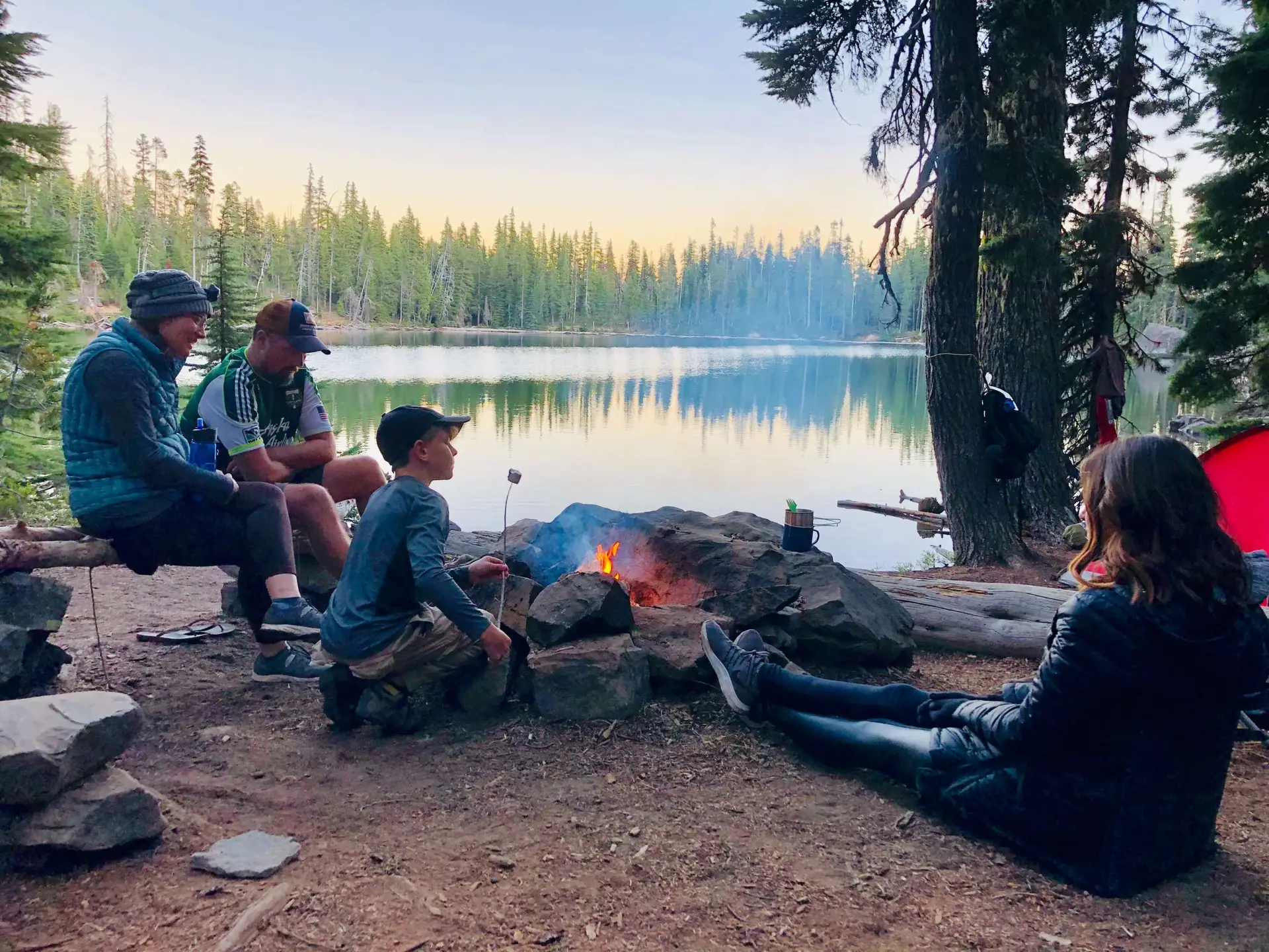 Four people gather around a campfire by a lakeside, surrounded by trees. One person is roasting marshmallows while sharing campfire stories for kids. Nearby, a red tent and backpacks sit under a serene sunset over the water.