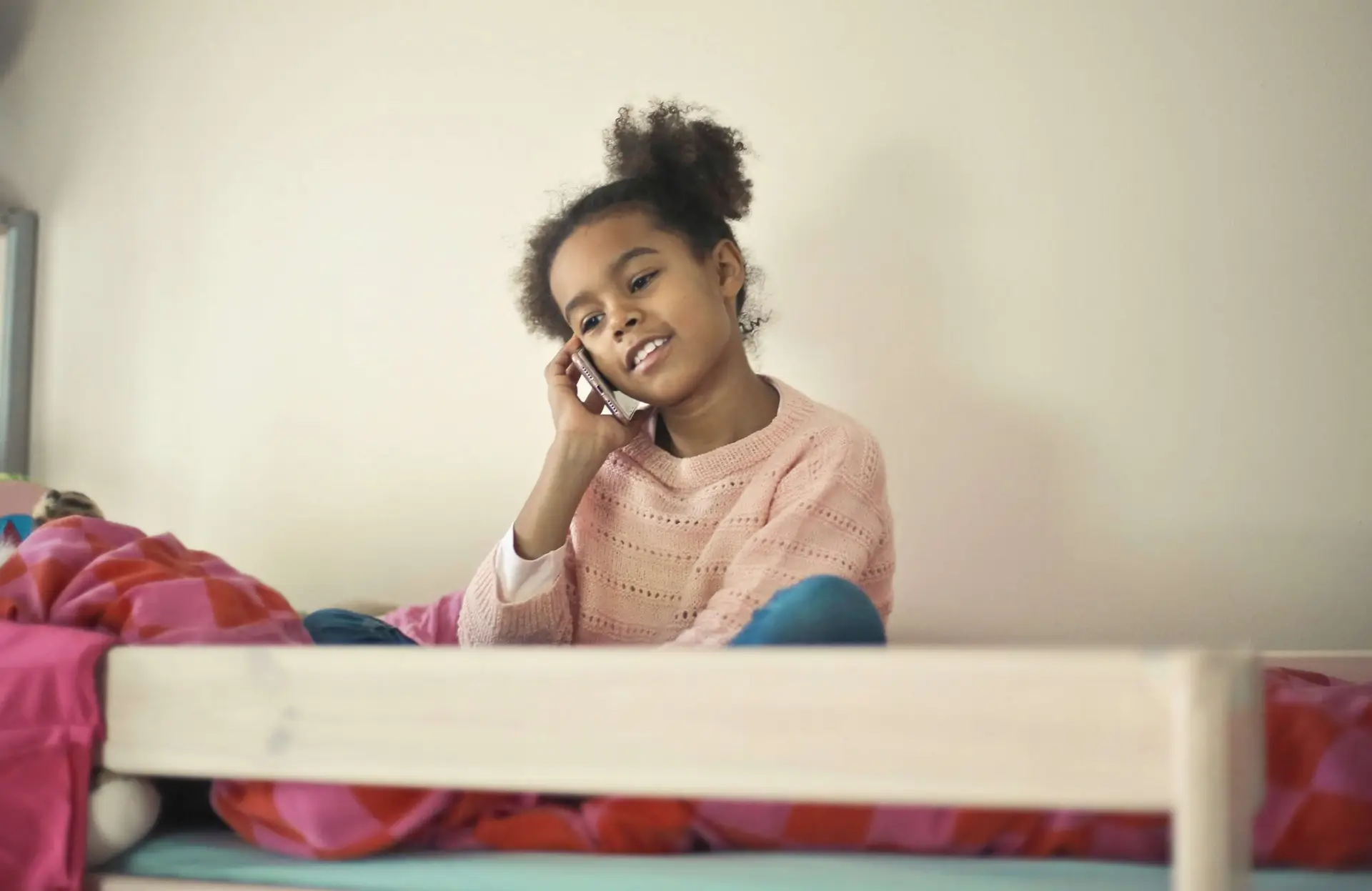 A girl with curly hair sits on a bed, smiling while discussing why kids should have phones in school. Dressed in a pink sweater, she is surrounded by colorful bedding in a room with a light, neutral background.