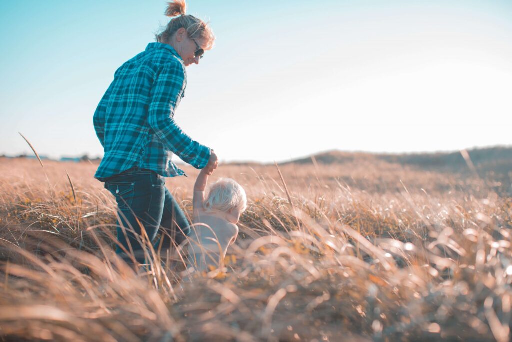 An adult in a blue plaid shirt holds hands with a small child, enjoying preschool fall activities as they walk through a sunlit field of tall grass. The sky is clear, and the scene is peaceful and serene.