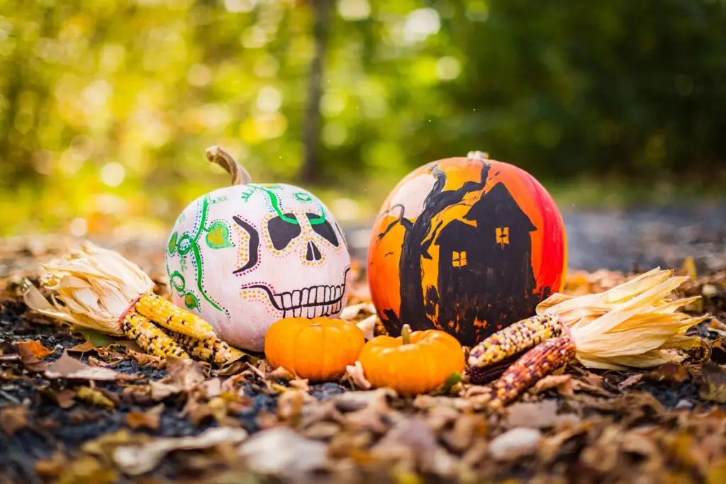 Two pumpkins are painted with Halloween designs: one with a skull and colorful patterns, the other with a haunted house silhouette. Scattered around them on the forest floor are mini pumpkins, ears of corn, autumn leaves, and whimsical popsicle stick crafts capturing the festive spirit.