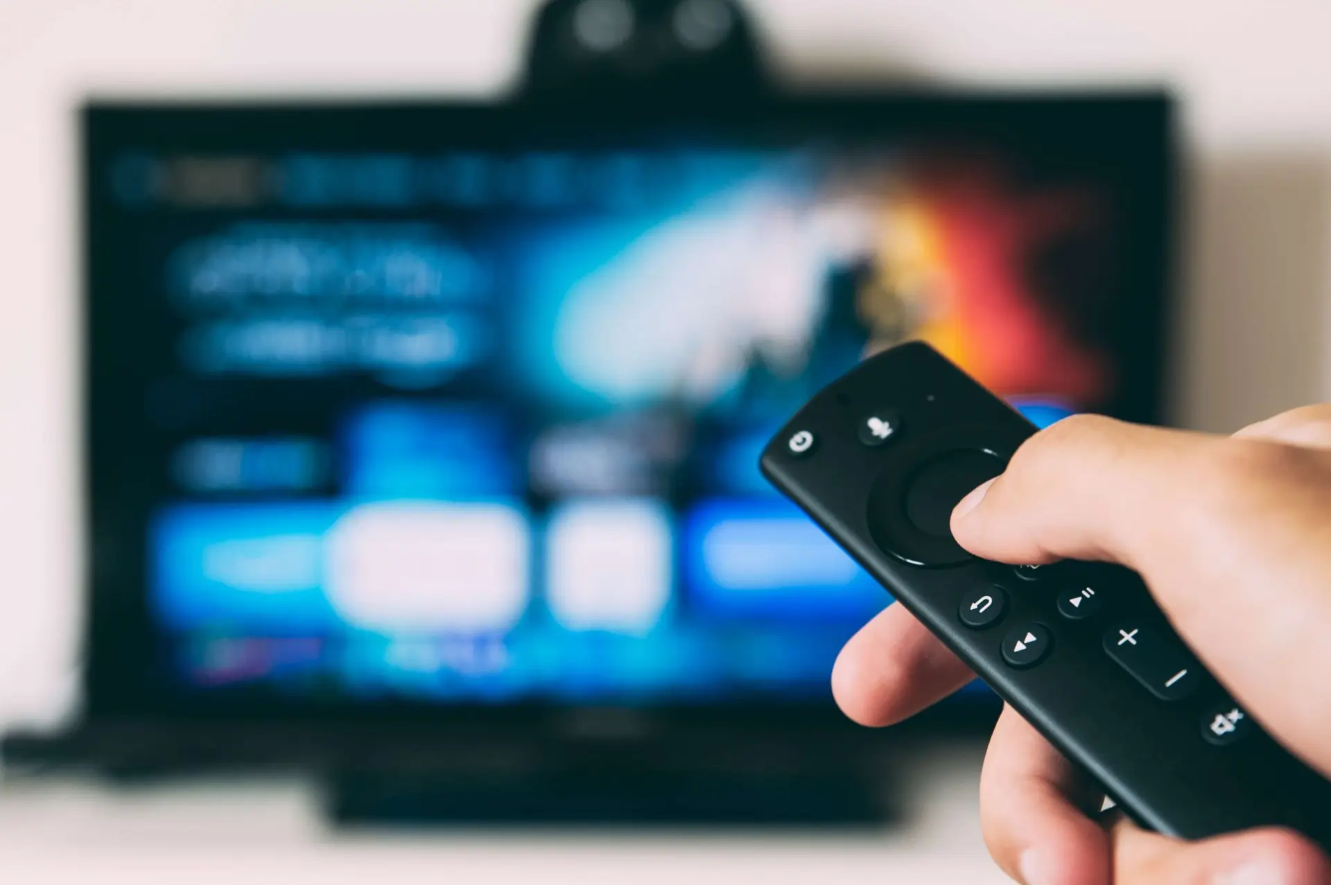 A hand holding a black remote control is pointed at a television screen, its blue and orange background blurred. Could this be the moment parents ponder, "Why is Prime not good for kids?" as they select suitable content?