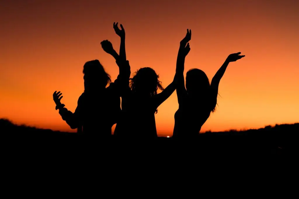 Silhouettes of three people dancing with raised arms against a vibrant orange sunset sky evoke the energy of minute to win it games for teens. The scene conveys a sense of joy and celebration.