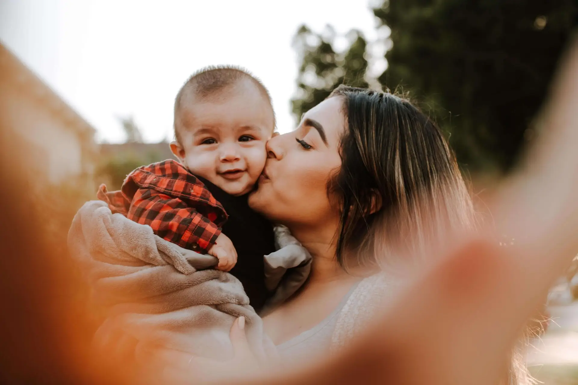 A woman gently kisses a smiling baby wrapped in a blanket, enjoying one of their favorite fall activities for infants. Surrounded by blurred greenery, the sun casts a warm, golden glow as she holds the baby close, creating a tender and affectionate moment outdoors.